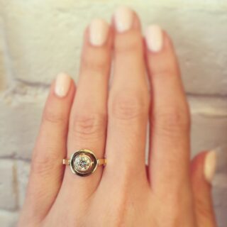 Close-up of a vintage‑inspired modernist solitaire ring with old European cut diamond set in milgrain bezel and golden crosshair prongs, on a flat gold hoop.