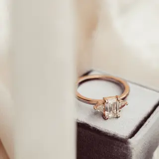 Close-up of emerald‑cut 1.0 ct colorless (DEF), VVS clarity moissanite with trillion‑cut side stones in a gold three‑stone engagement ring