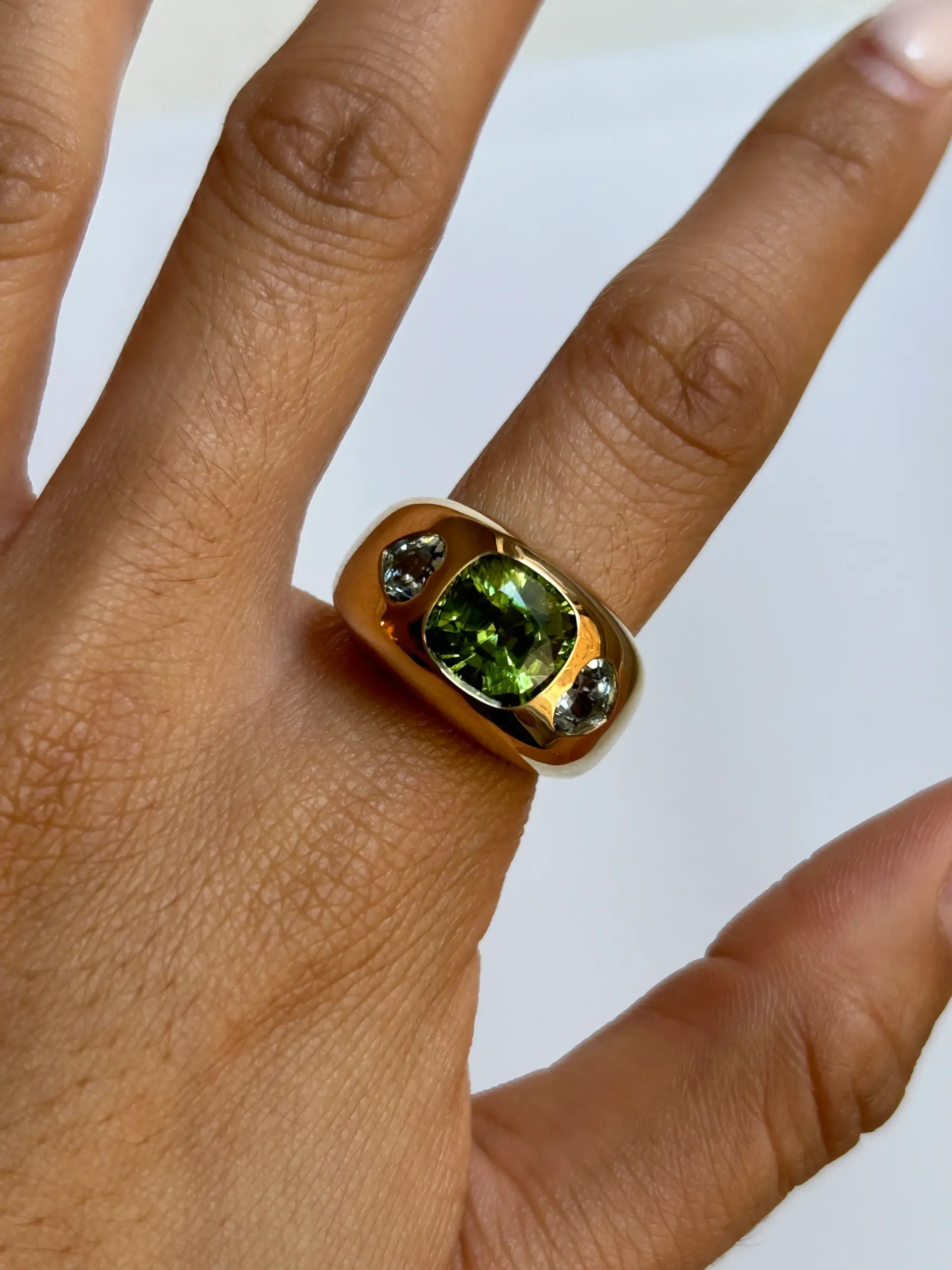 Angled shot showing the ring’s 14 mm gold band and heart-shaped side aquamarine gemstones flanking the cushion-cut peridot.