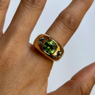 Angled shot showing the ring’s 14 mm gold band and heart-shaped side aquamarine gemstones flanking the cushion-cut peridot.