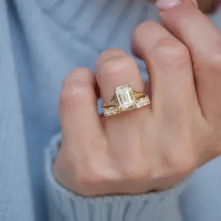 Berlinger split‑shank solitaire ring held between fingers, showcasing the emerald‑cut diamond’s cut clarity and detailed split shank band in warm yellow gold