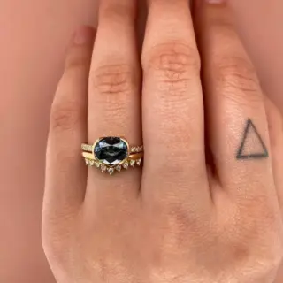 Close-up of the Estrada Ring showing a dramatic 2.31 ct dark gray‑blue spinel set horizontally (east‑west) in a 14k gold half‑bezel, accented by a half band of .09 ctw SI G‑H diamonds