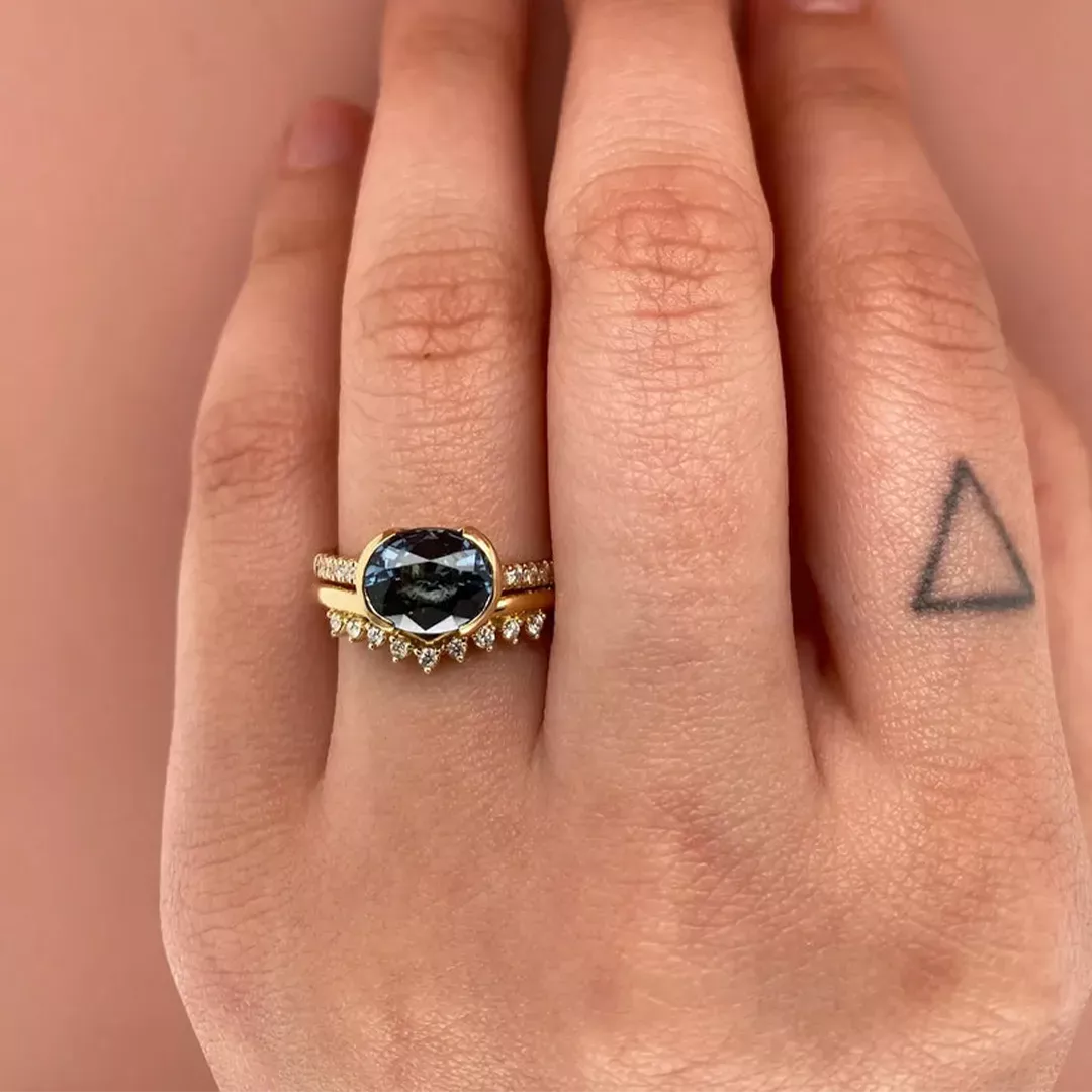 Close-up of the Estrada Ring showing a dramatic 2.31 ct dark gray‑blue spinel set horizontally (east‑west) in a 14k gold half‑bezel, accented by a half band of .09 ctw SI G‑H diamonds