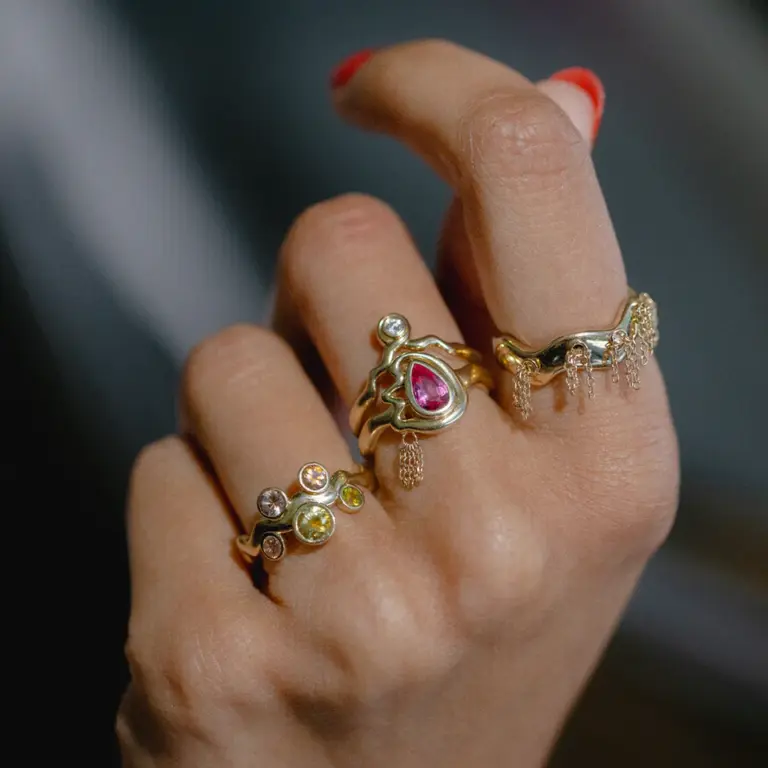 Close-up of pink sapphire ring with sculptural bezel setting and delicate gold tassel chains on a polished gold band.